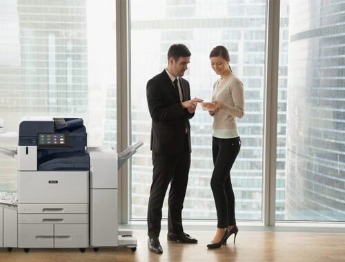 Twee mensen in een kantoor in zakelijke kleding die documenten bespreken, staan ​​bij een Xerox® AltaLink® B8200 Series Multifunctionele Printer